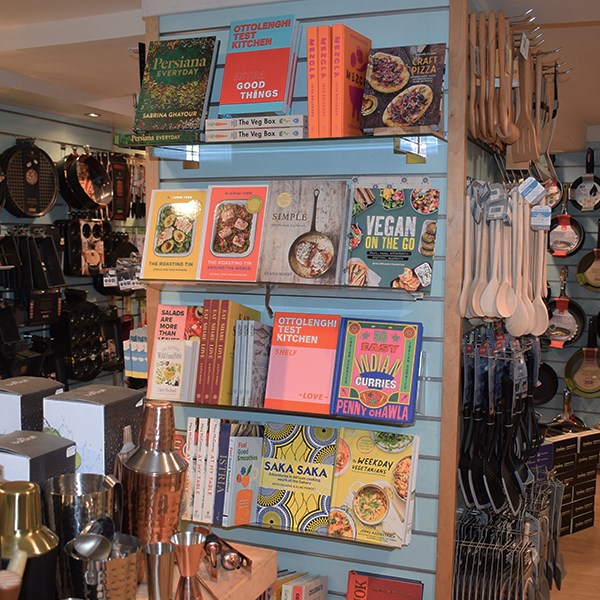 Cookbooks on shelving in a shop setting selling spoons, pans and cocktail shakers.