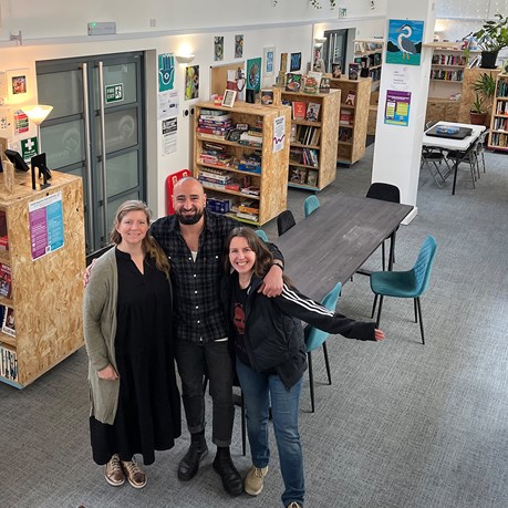 Three people standing together in a library space, while smiling and looking at camera.