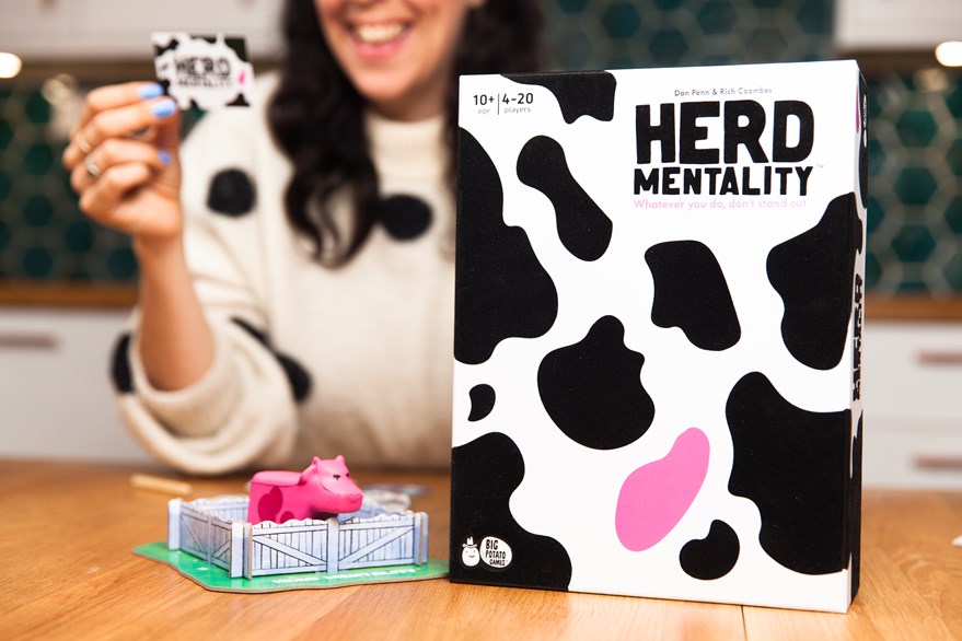 Cow print Herd Mentality game standing upright on a table with a woman in the background reading a card.