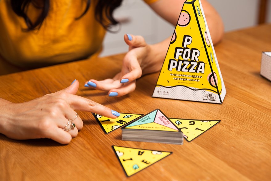 P for Pizza game in a yellow triangular box on a table with a woman's hand pointing at the cards from the game.